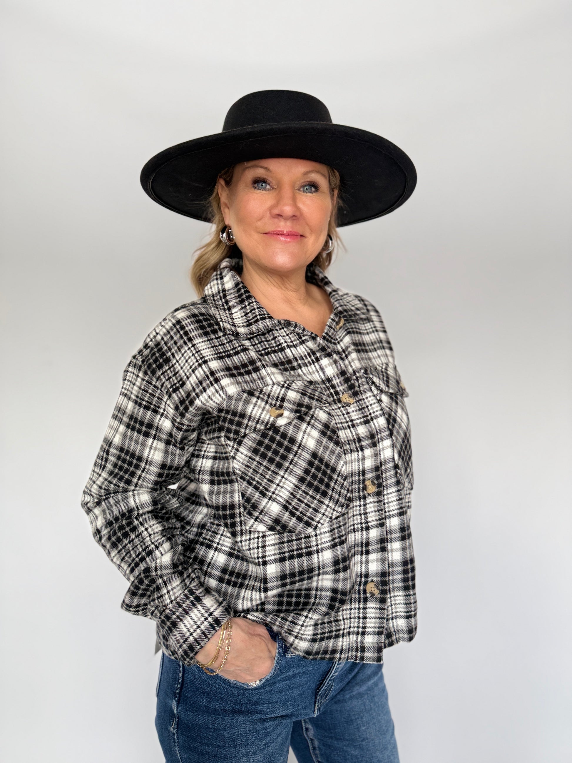 Person wearing a black and white plaid shirt, blue jeans, and a black hat on a plain background