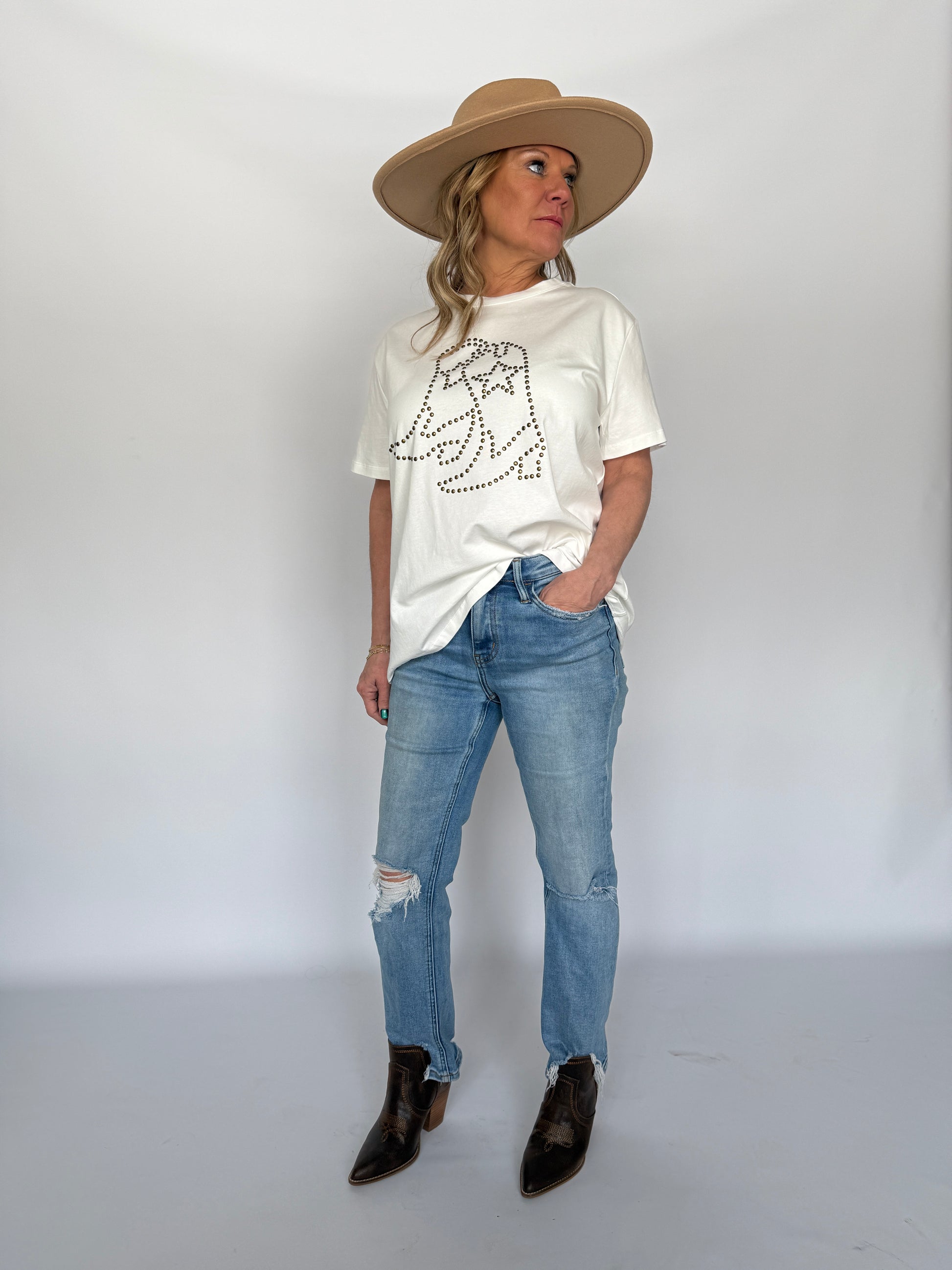 Person wearing a white t-shirt with a graphic design, blue jeans, and a beige hat on a plain background